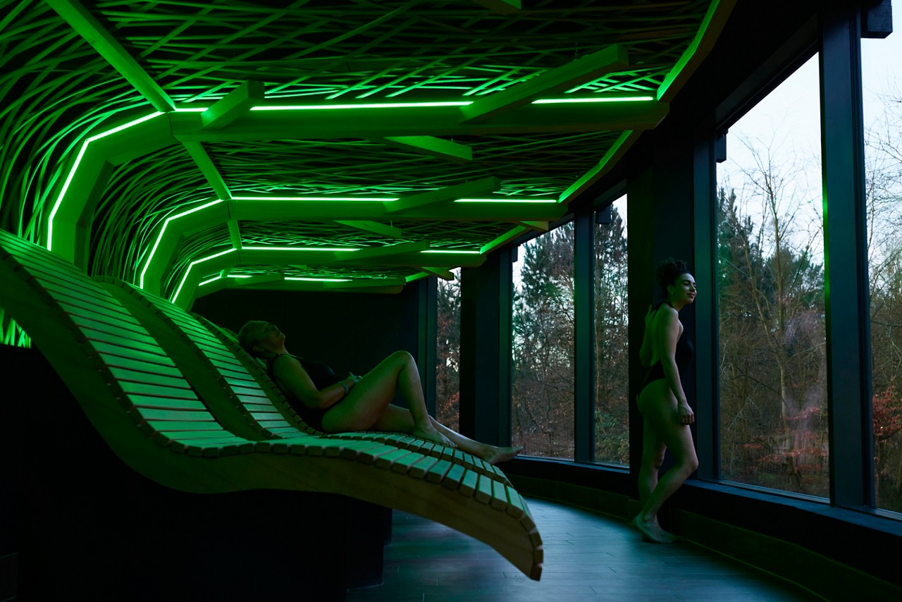 Two people in swimsuits relax—one reclining on curved wooden loungers, one standing and gazing outside—inside a spa room with glowing green geometric ceiling and tall windows overlooking trees.